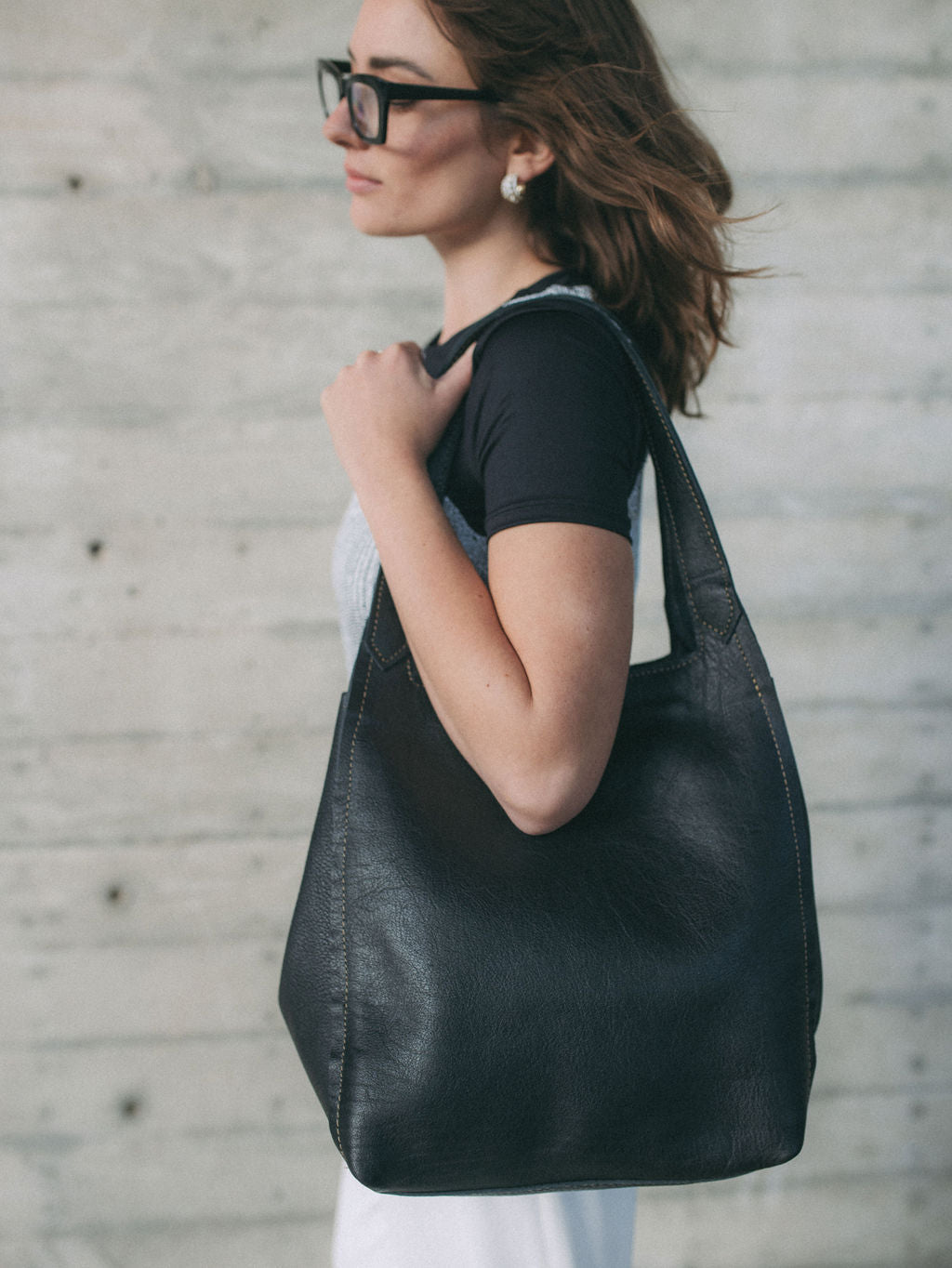 A stylish woman walking in a city, wearing a modern outfit and carrying a sleek black luxury leather shoulder bag. The bag is shown draped casually over her shoulder.