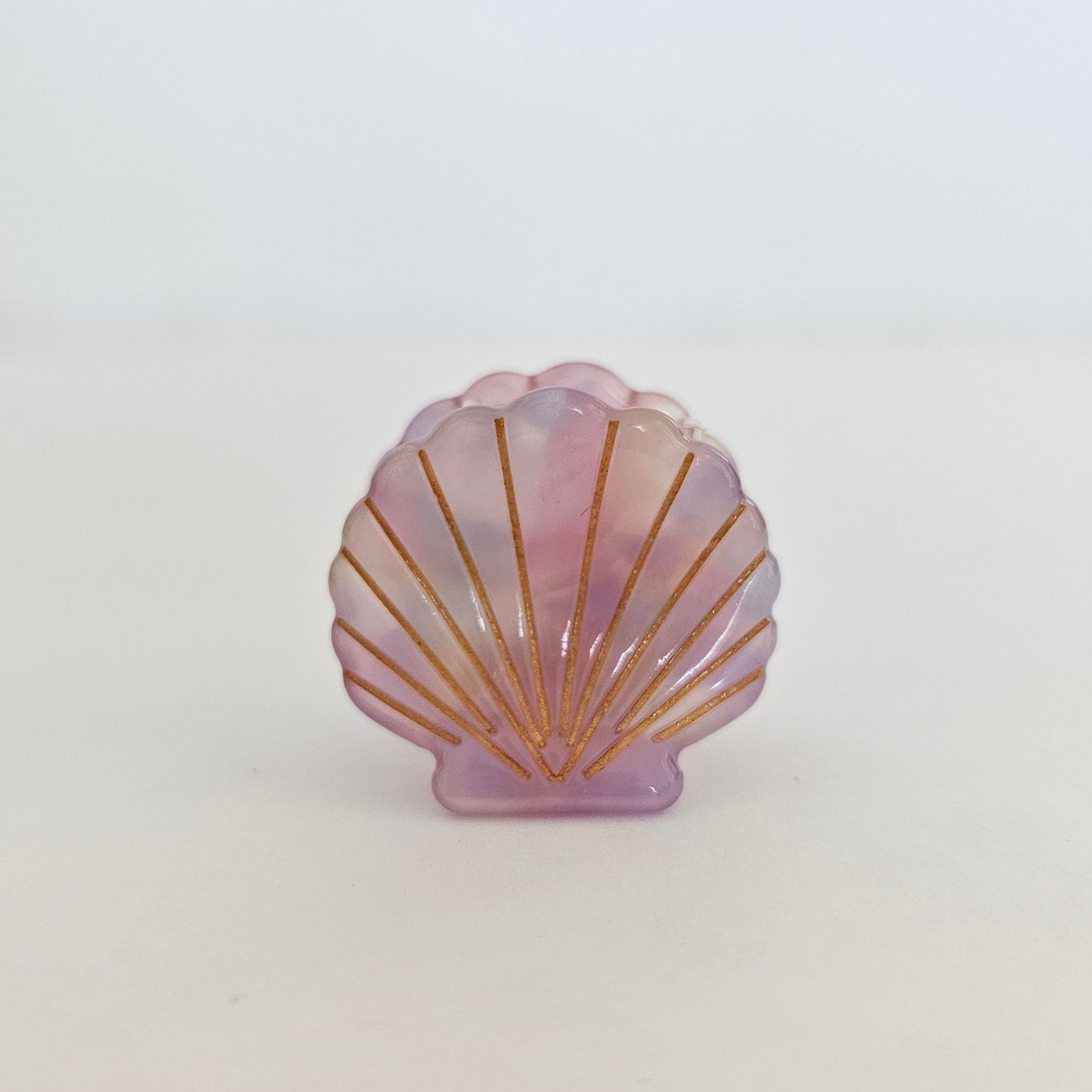 A small shell-shaped decorative object with a translucent pink and lavender iridescent finish and gold-toned radial lines, standing on a white surface against a white background.